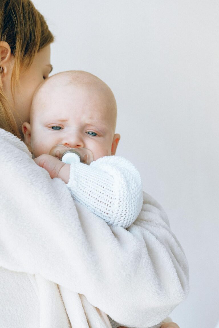 Side view of casual female giving love and hug to cute newborn baby in knitted clothes while standing together in bedroom